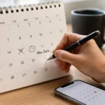 Tangan mencoret tanggal lama dan melingkari tanggal baru di kalender meja menggambarkan makna reschedule artinya menjadwalkan ulang